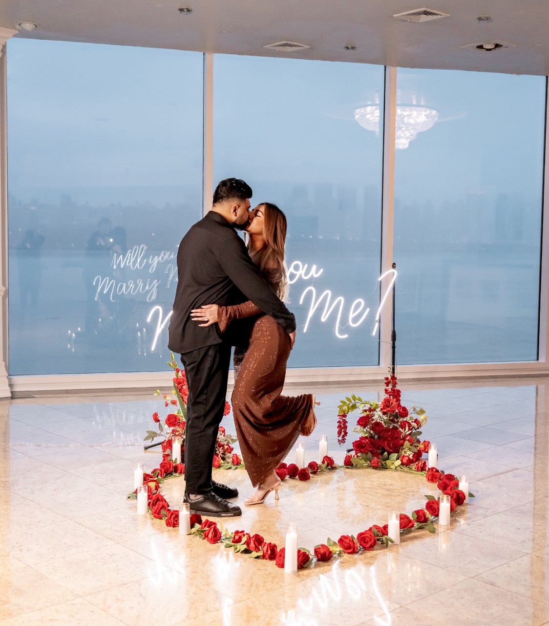 Couple embracing inside a glowing 'Marry Me' neon sign display with rose petals on a high-floor room with the New York City skyline behind them
