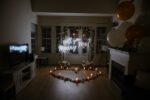Indoor living room proposal setup with 'Will You Marry Me' neon sign, gold and white balloons, and candle-lit heart arrangement